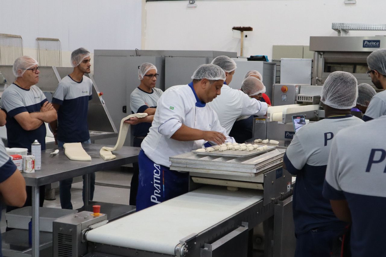 Um grupo de homens está em pé ao redor de uma esteira rolante em uma padaria.