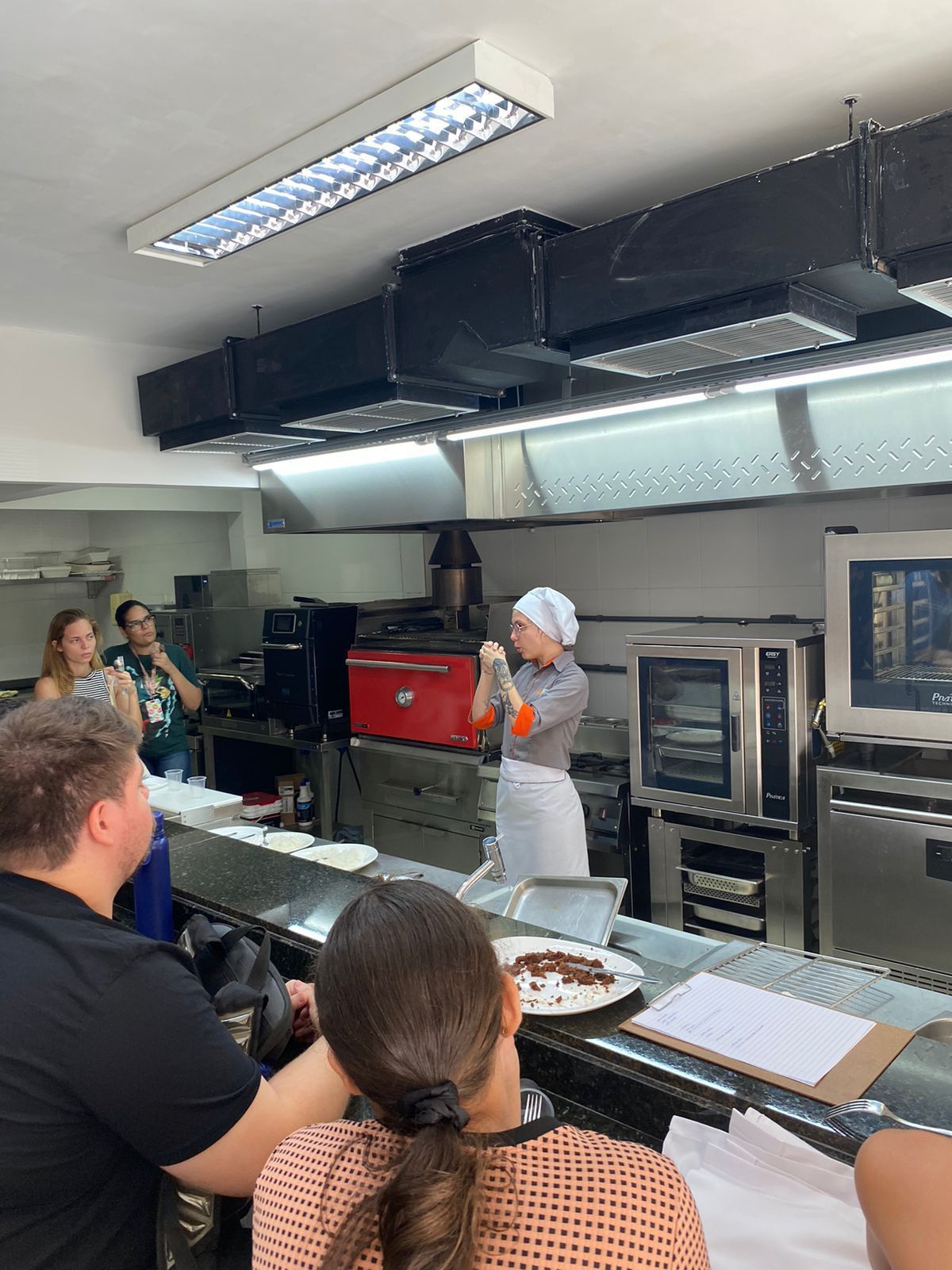 Um grupo de pessoas está sentado à mesa na cozinha observando uma mulher cozinhar.