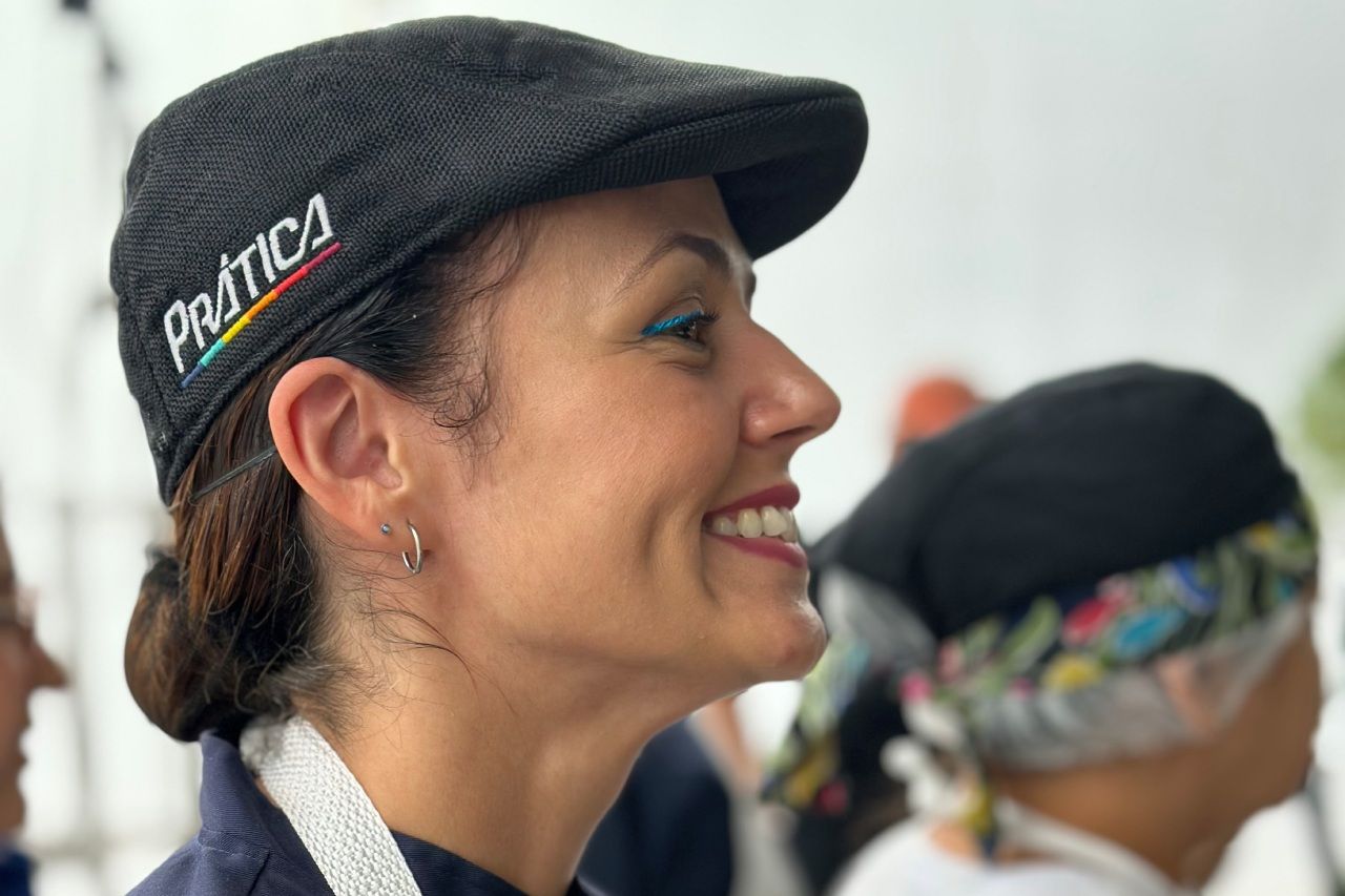 Mulher sorrindo, usando um boné preto com a palavra “Política” estampada. Ela está em um ambiente externo com outra pessoa ao fundo.