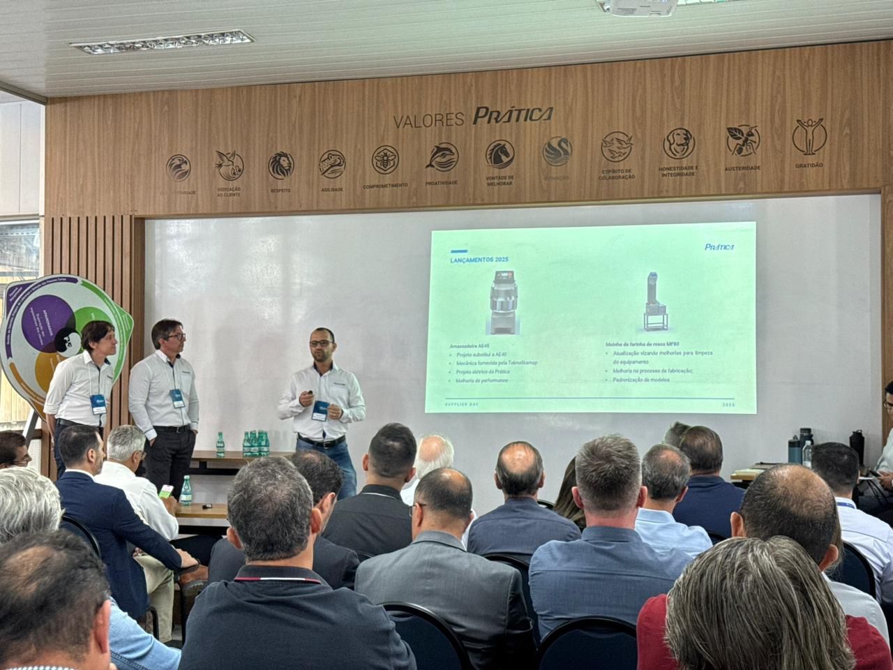 Uma apresentação em uma sala de conferências com um palestrante e plateia. Dois homens em pé à esquerda do palestrante.