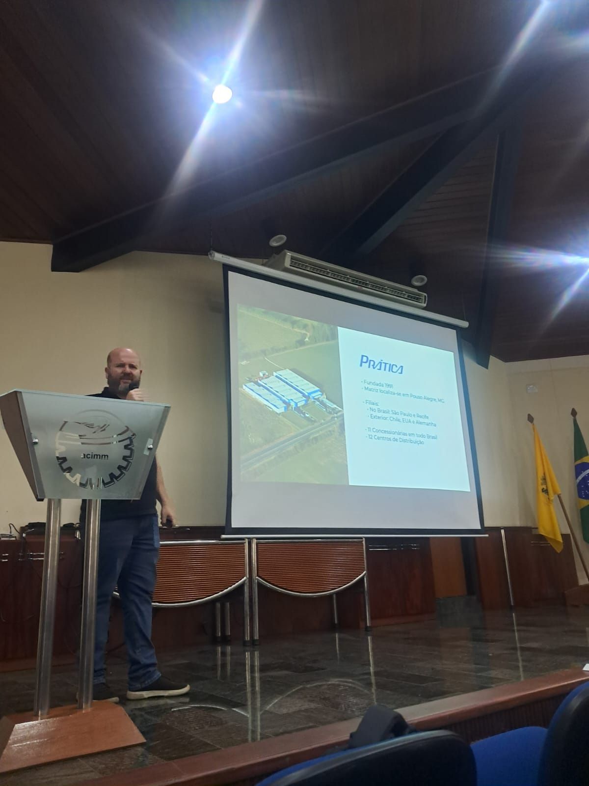 Homem apresentando no pódio, slides mostrando o layout das instalações. Sala marrom, bandeiras à direita.