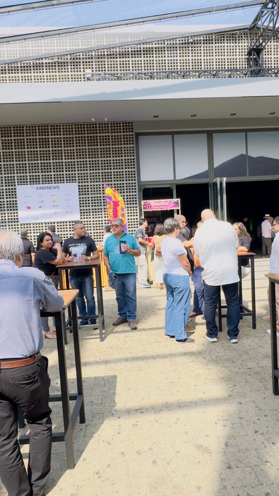 Um grupo de pessoas está de pé ao redor de mesas do lado de fora de um prédio.