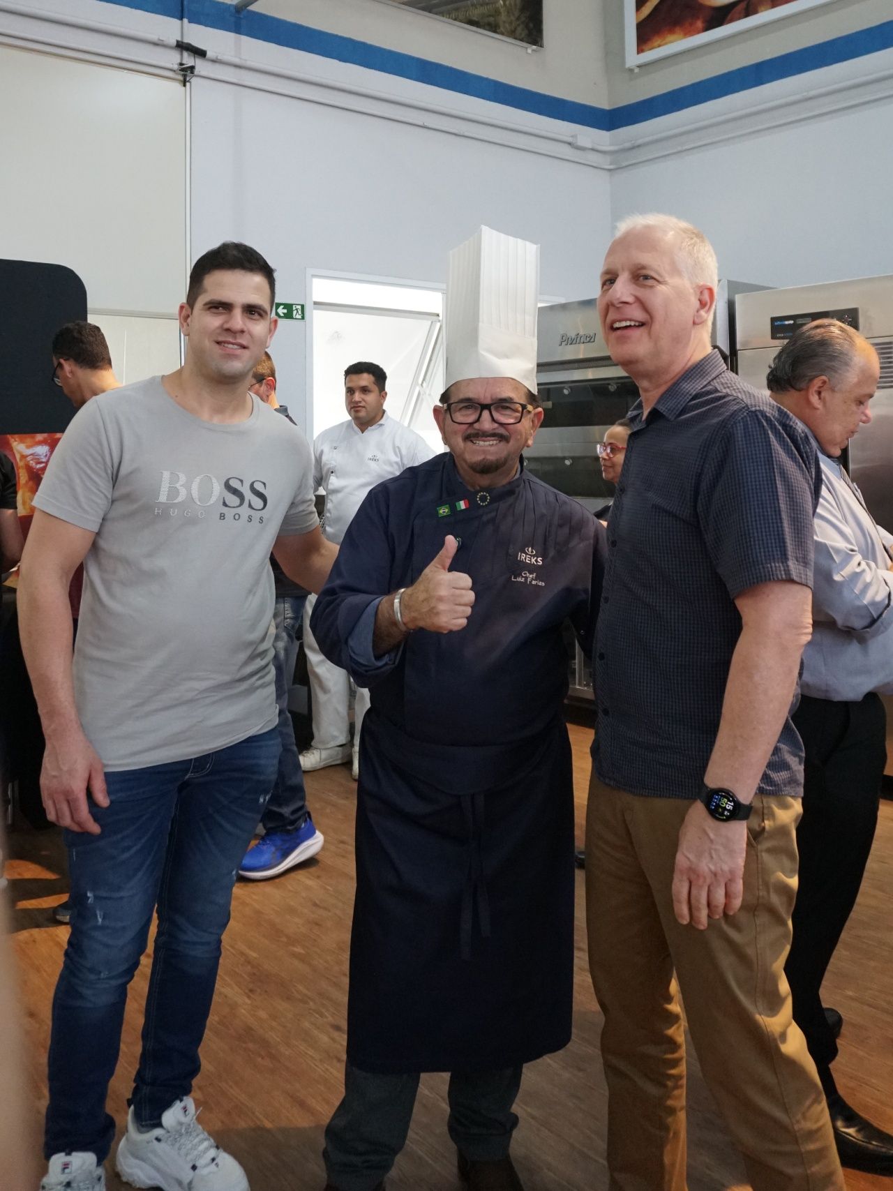 Três homens posam: um chef uniformizado faz sinal de positivo, outros sorriem em uma sala com piso de madeira.