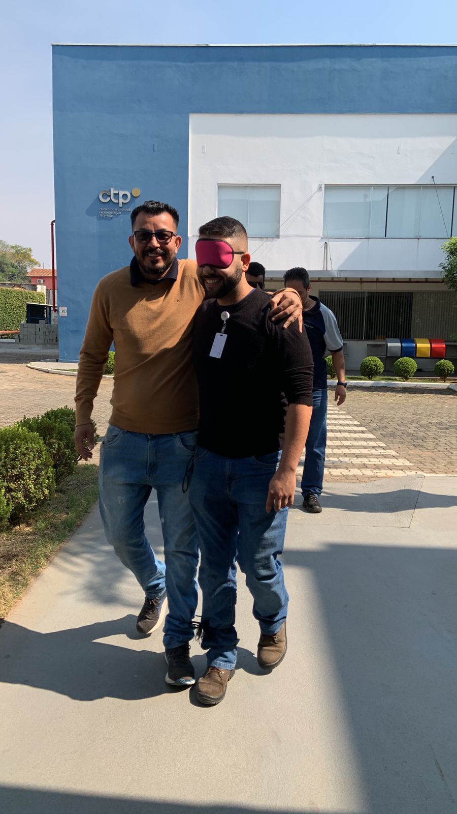 Dois homens estão caminhando pela calçada, um deles usando uma venda nos olhos.