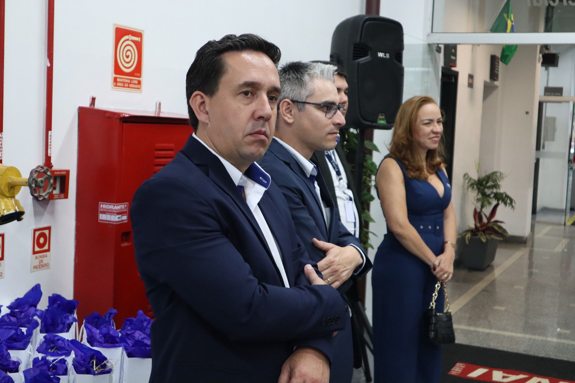 Três pessoas em pé dentro de um prédio. Homem de blazer azul-marinho, dois homens e mulher de vestido azul.