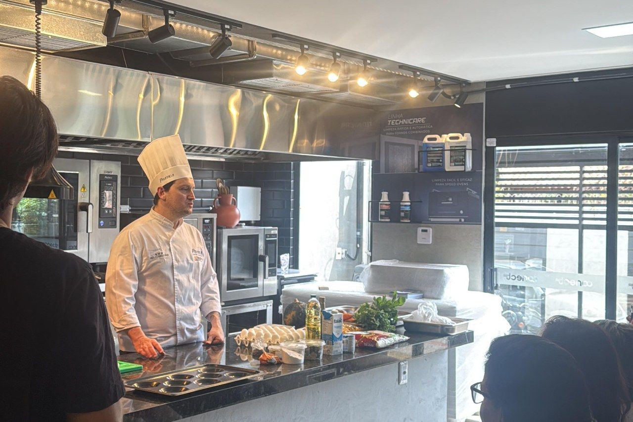 Chef de branco na cozinha; dando uma palestra em um balcão; alguns ingredientes crus expostos à sua frente.