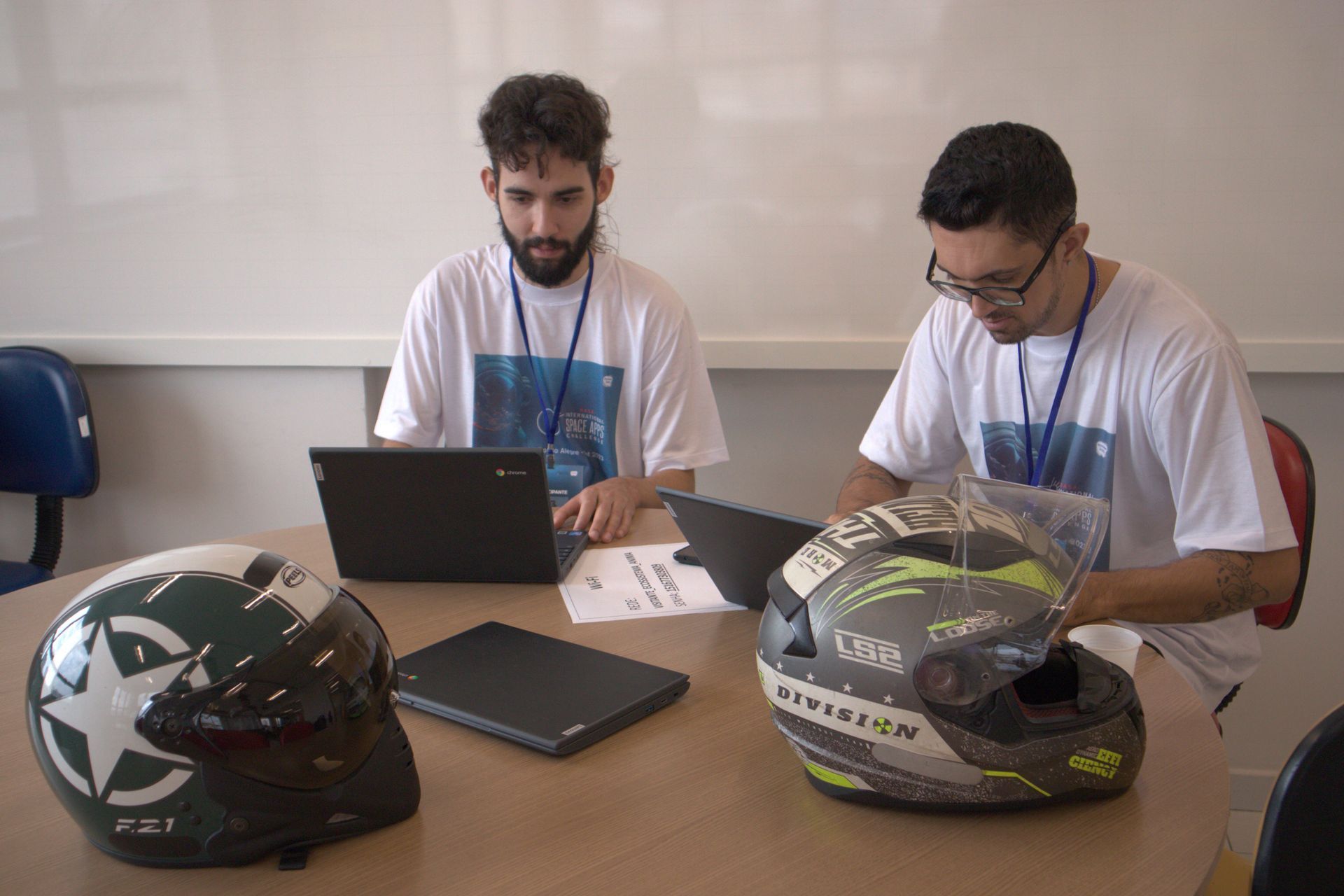 Dois homens estão sentados à mesa com capacetes e um laptop.
