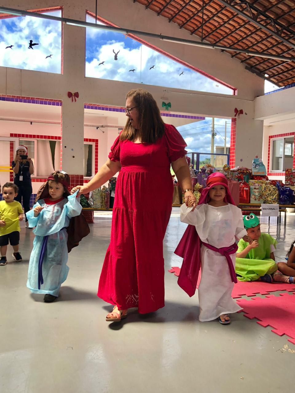 Uma mulher de vestido vermelho está de mãos dadas com duas crianças fantasiadas.