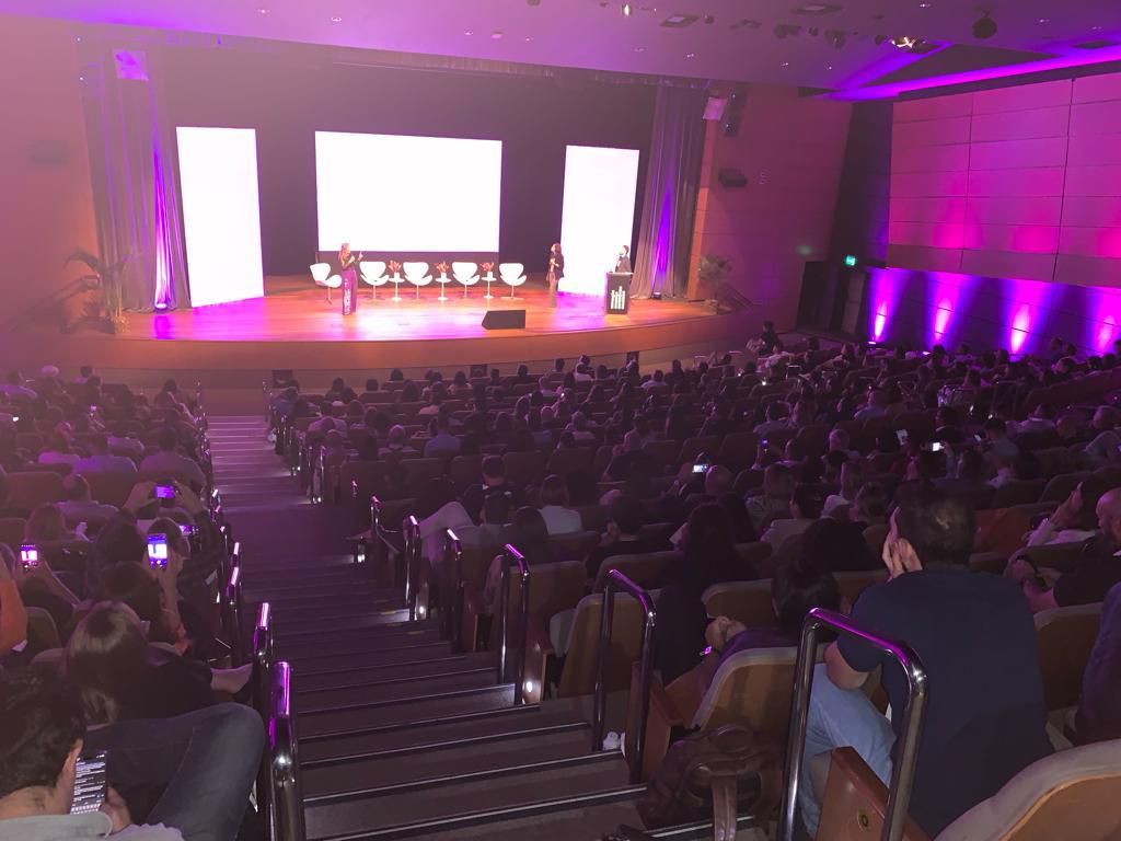 Uma grande multidão de pessoas está sentada em um auditório assistindo a uma apresentação.