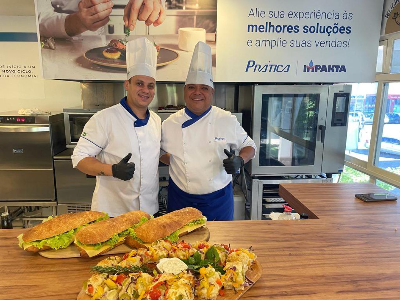 Dois chefs estão de pé, um ao lado do outro, em uma cozinha.