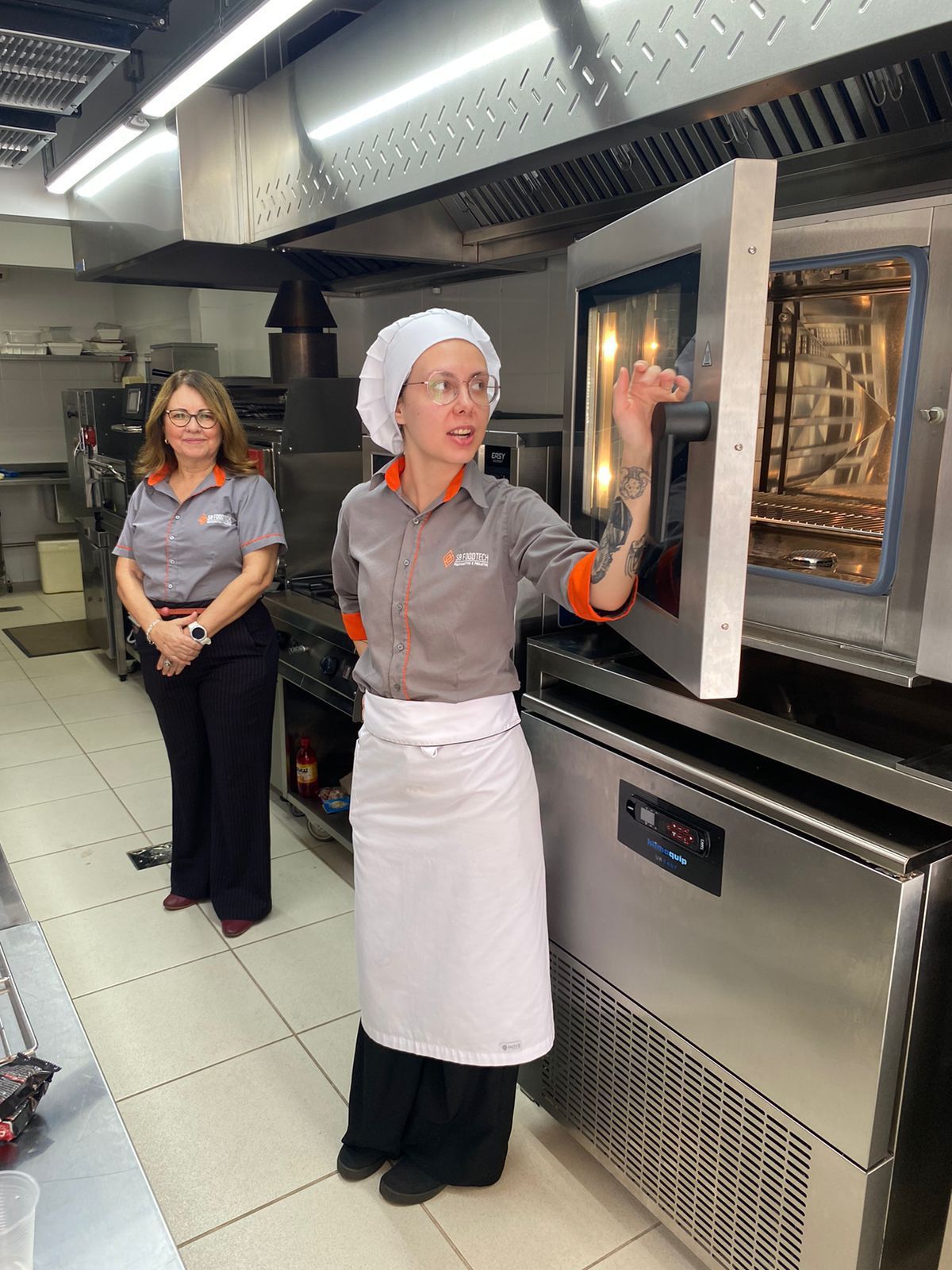 Uma mulher está abrindo um micro-ondas na cozinha enquanto outra mulher está atrás dela.