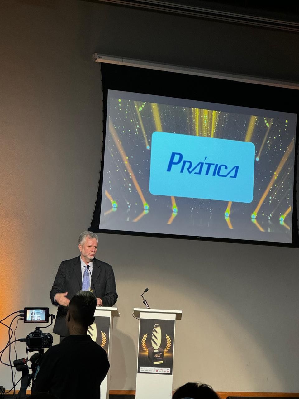 Um homem está em um pódio em frente a uma tela que diz pratica