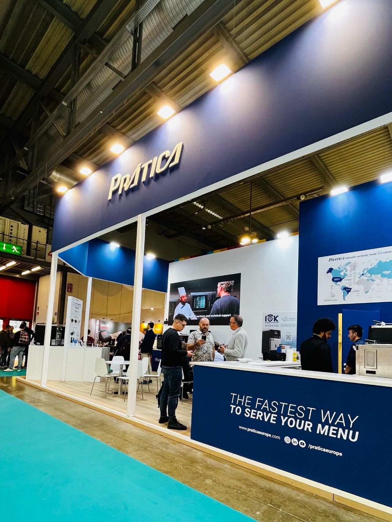 Estande azul e branco da Pratika em uma feira. Pessoas reunidas ao redor do balcão e dentro do estande. Texto: 
