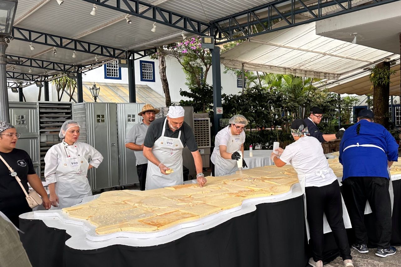Pessoas preparando uma massa folhada muito comprida em uma mesa ao ar livre, sob um toldo de metal.