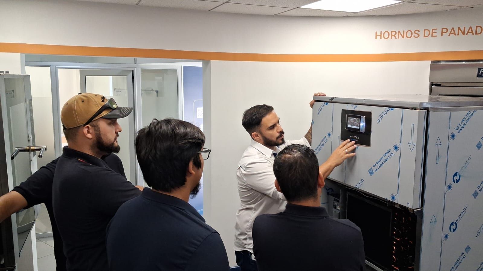 Quatro homens examinam um painel de controle de um forno industrial na sala de equipamentos de uma padaria.