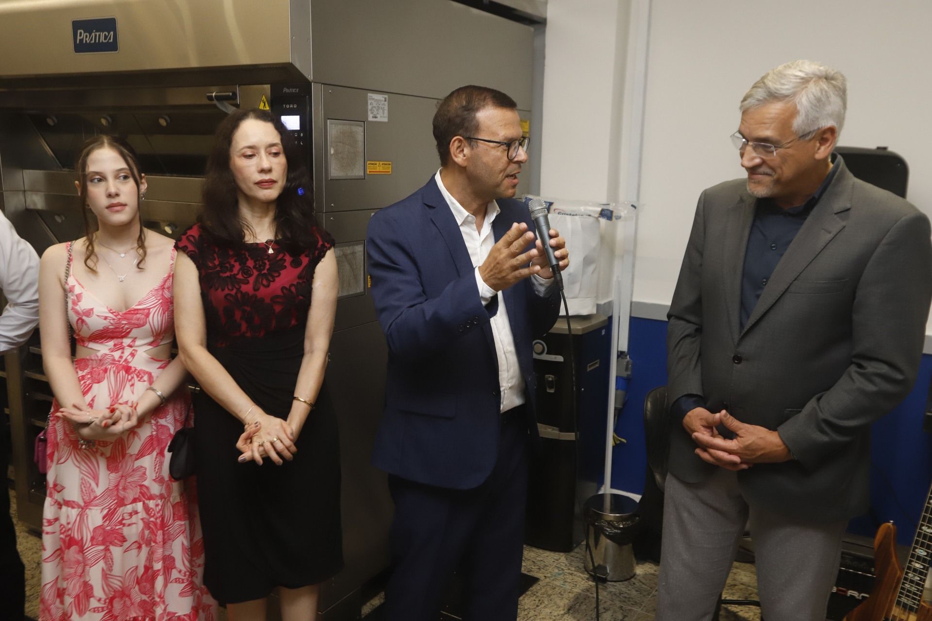 Quatro pessoas em um evento, um homem falando em um microfone, outros ouvindo, em uma cozinha.