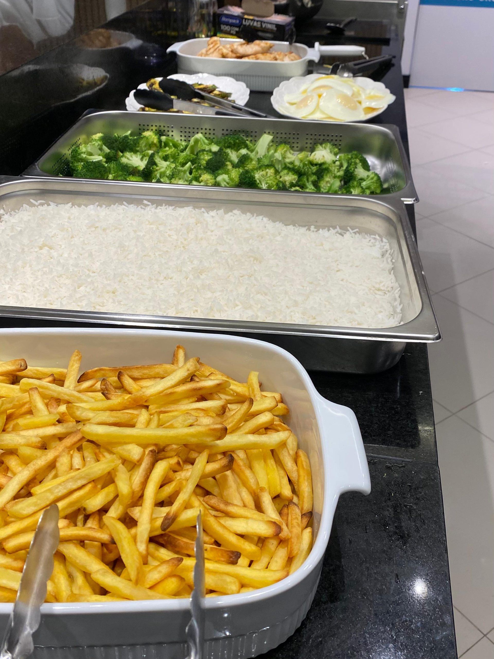 Uma bandeja de batatas fritas fica em um balcão ao lado de uma bandeja de brócolis e arroz
