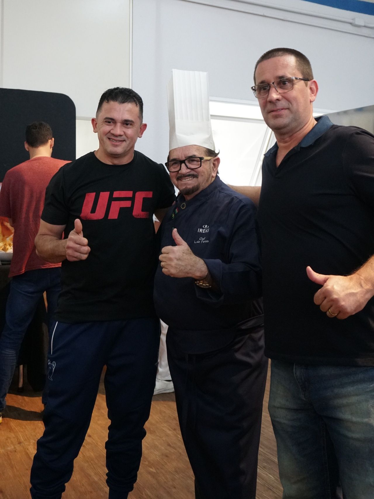 Três homens fazendo sinal de positivo. Um lutador de UFC com um chef de chapéu branco; outro homem de calça jeans azul.
