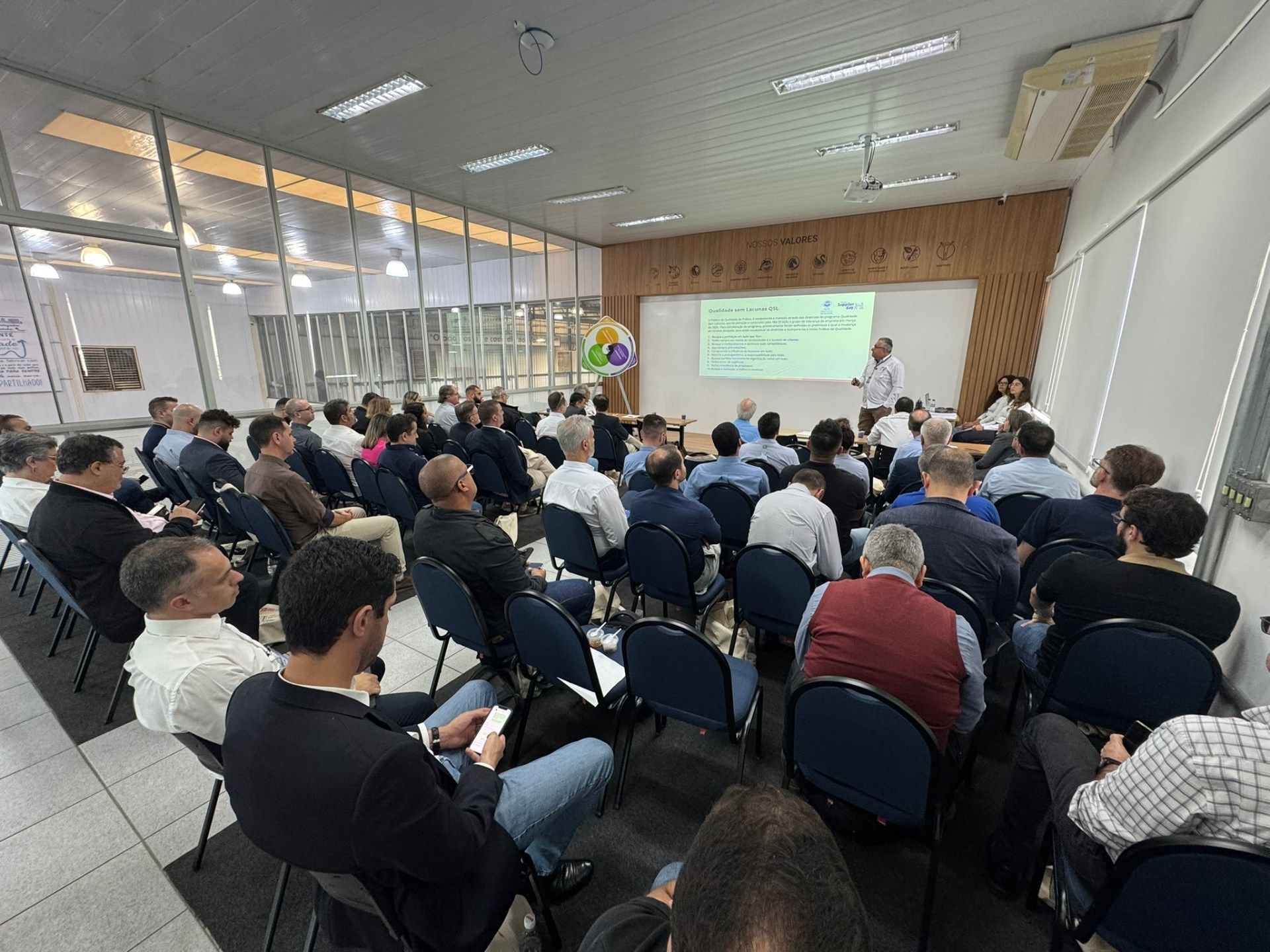 Um grupo de pessoas está sentado em cadeiras em uma sala assistindo a uma apresentação.