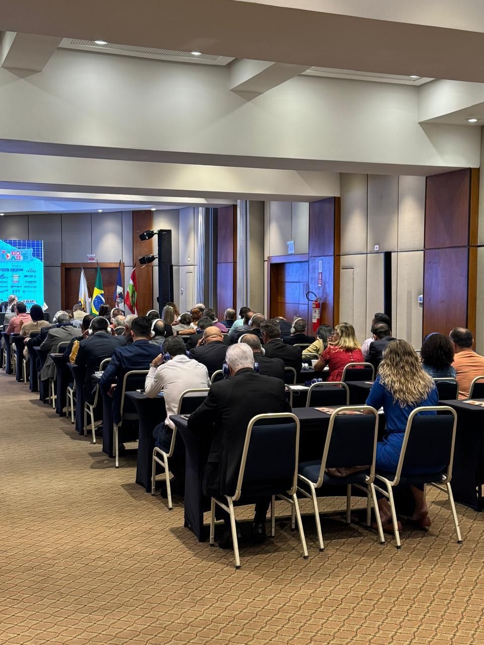 Um grande grupo de pessoas está sentado em mesas em uma sala de conferências.