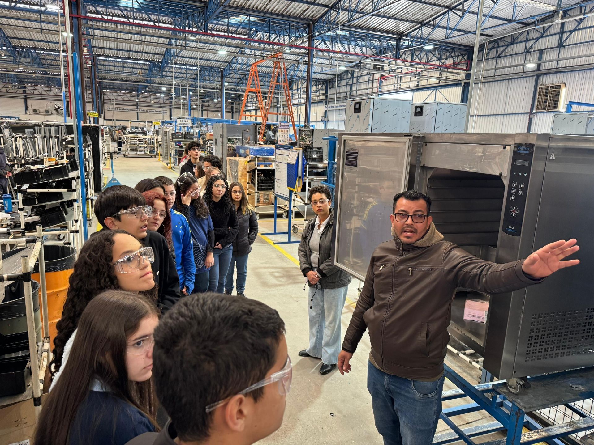 Grupo de estudantes em visita a uma fábrica, ouvindo um homem apontando para um equipamento.