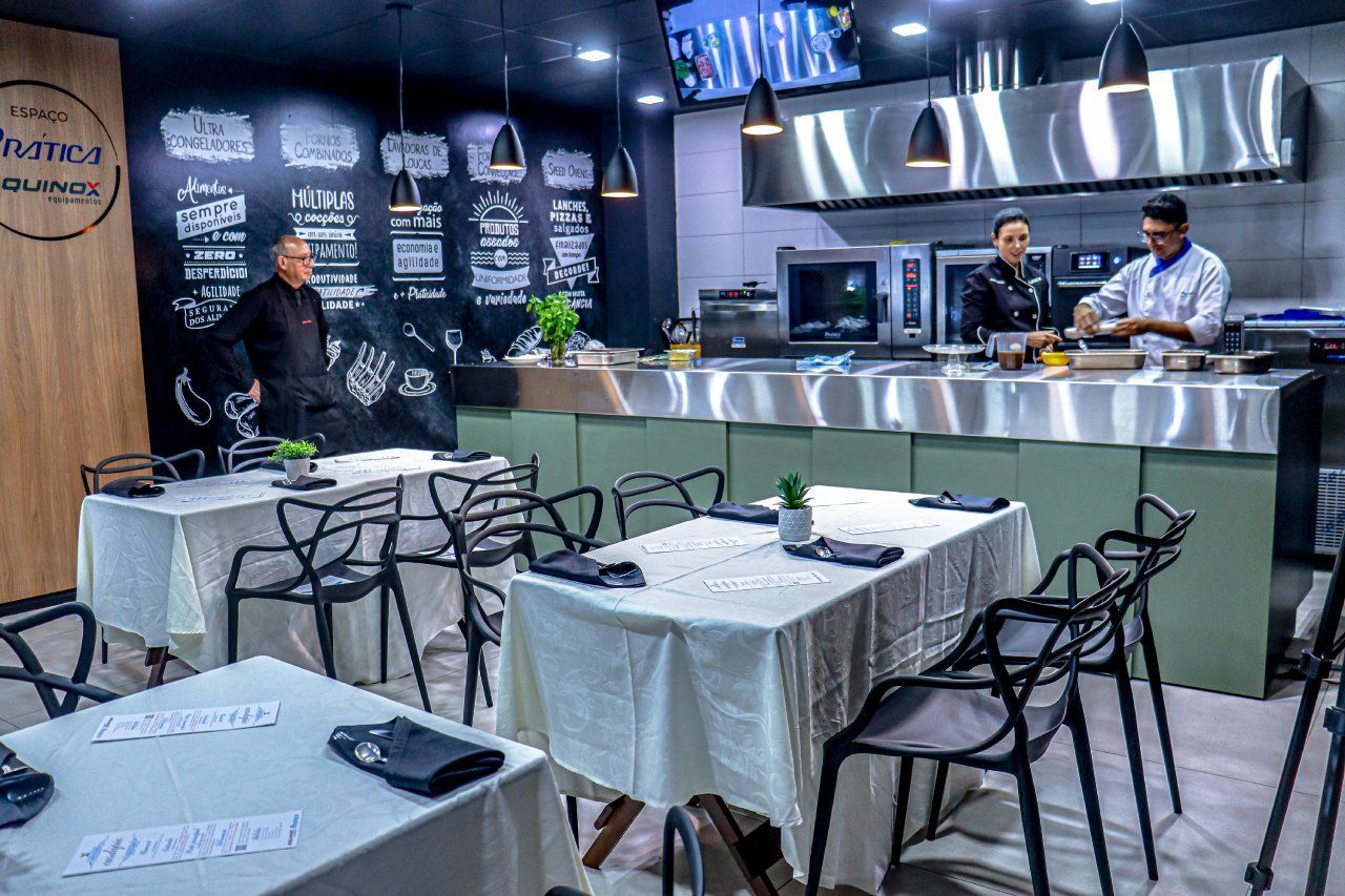 Um restaurante com mesas e cadeiras e um chef preparando comida ao fundo.