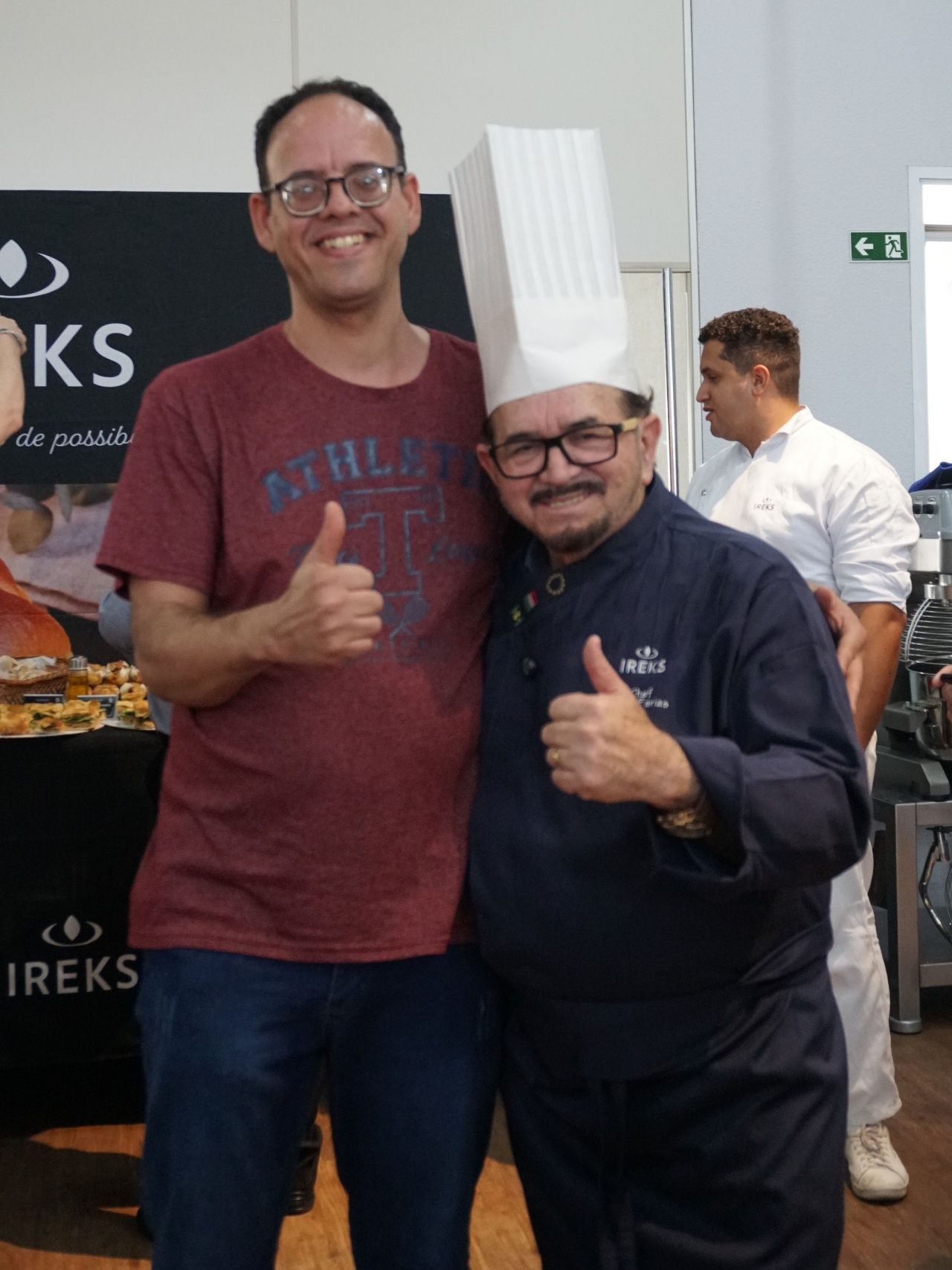 Homem de óculos faz sinal de positivo ao lado de um chef de chapéu branco, ambos sorrindo.
