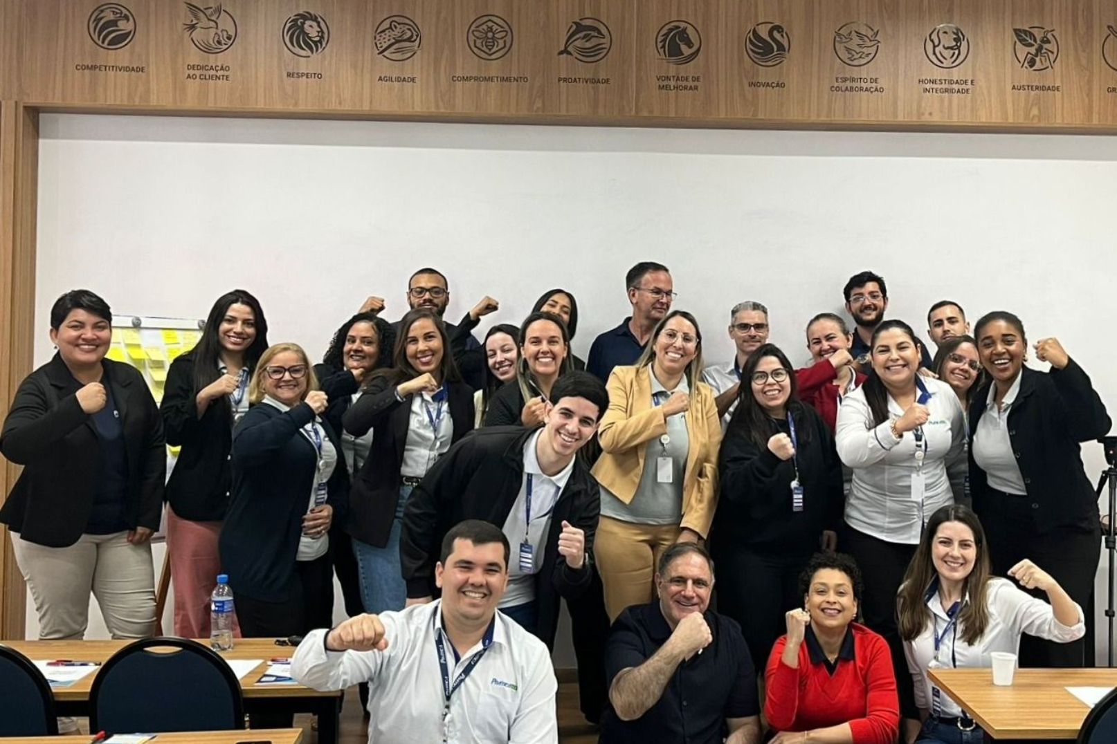 Grupo de pessoas sorrindo e flexionando os braços em uma sala de conferências bem iluminada.