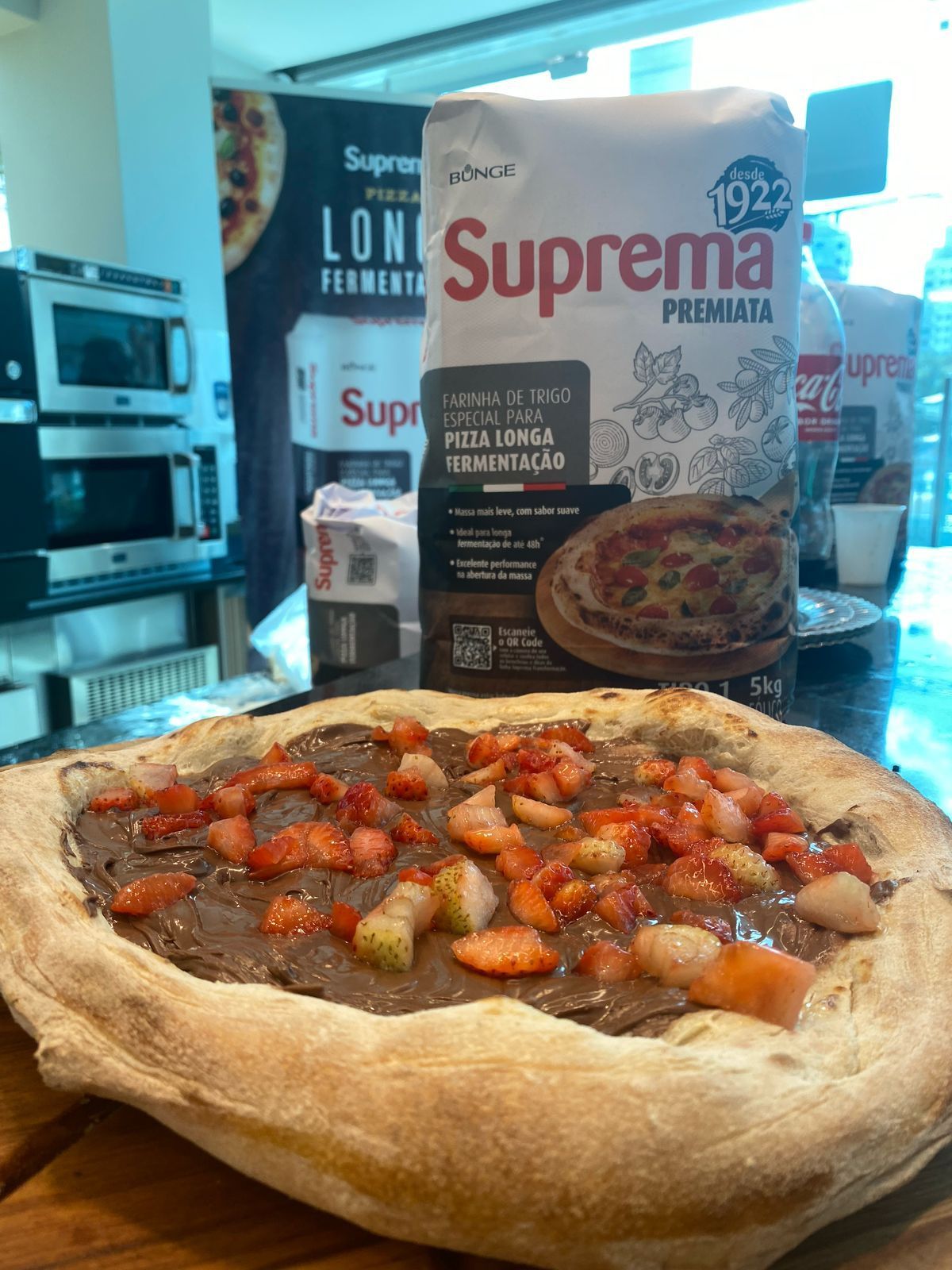 Uma pizza com morangos e calda de chocolate está sobre uma mesa de madeira ao lado de um saco de farinha suprema.