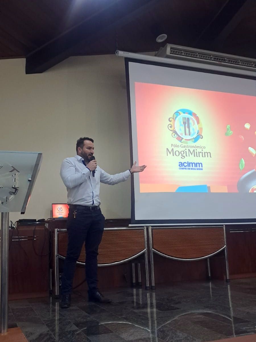 Homem de camisa azul claro, apresentando-se no pódio, gesticulando em direção a uma tela com o logotipo