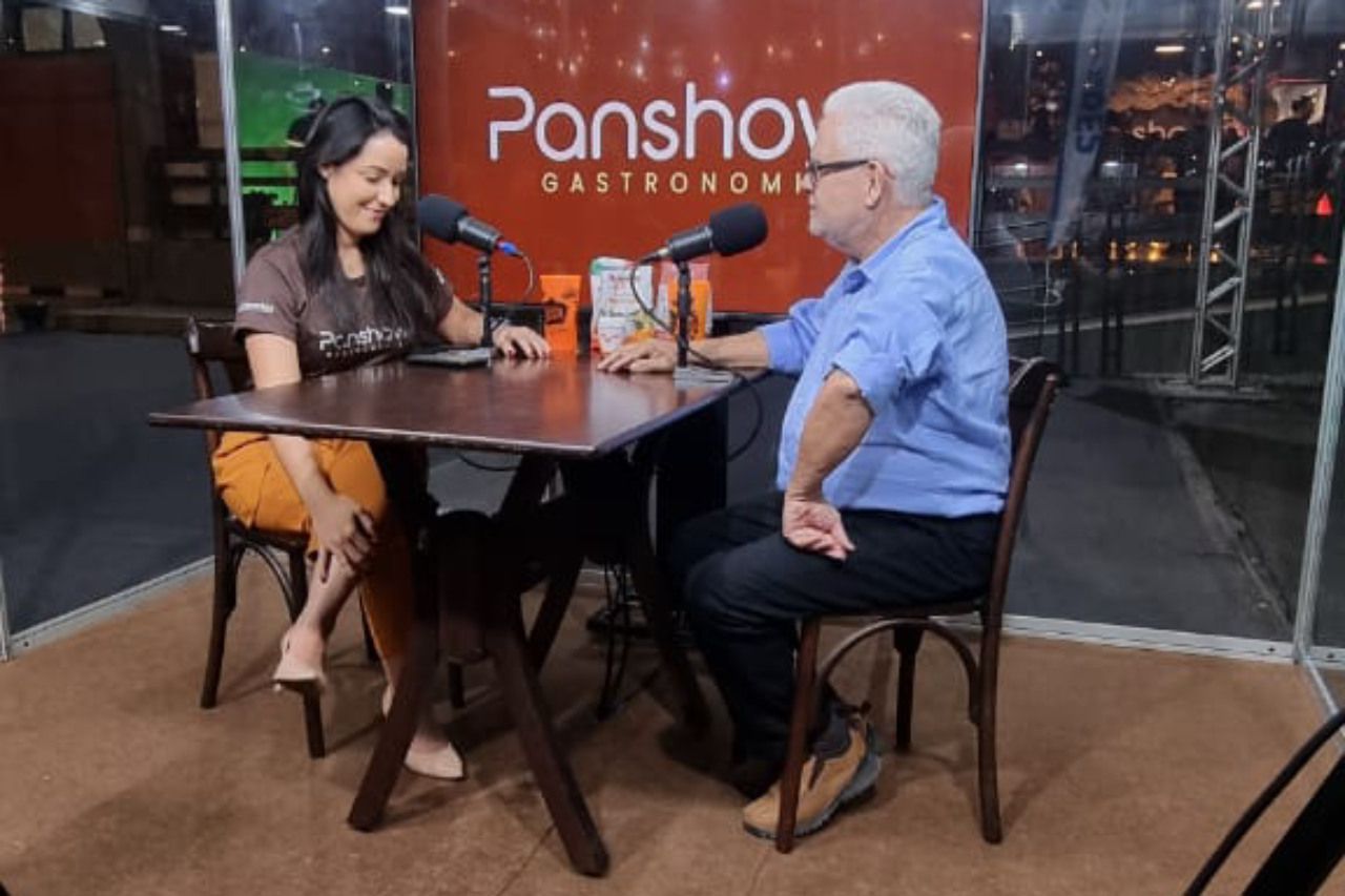 Um homem e uma mulher estão sentados em uma mesa com microfones em frente a uma placa do panshow