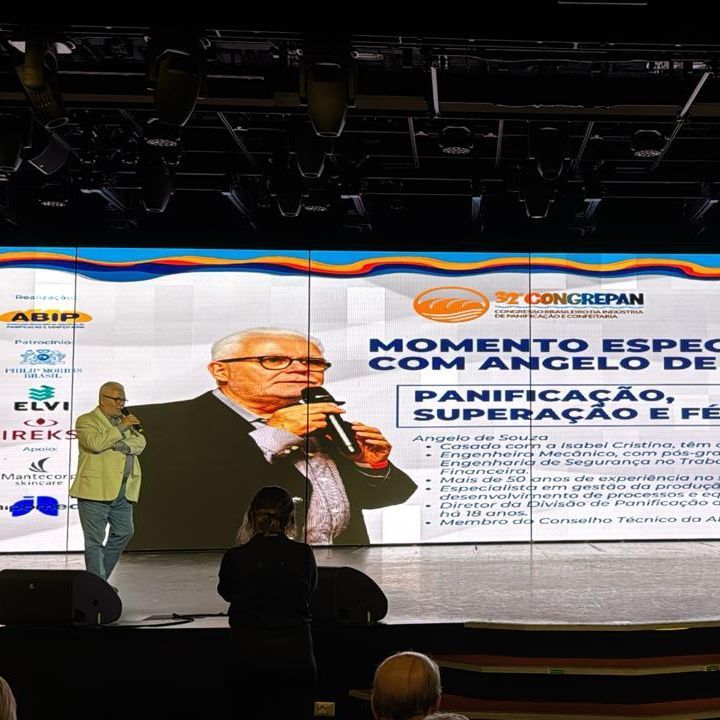 Um homem está em pé no palco segurando um microfone em frente a uma tela grande.