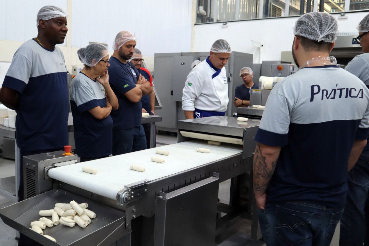 Um grupo de pessoas em pé ao redor de uma esteira rolante com um homem vestindo uma camisa que diz prática