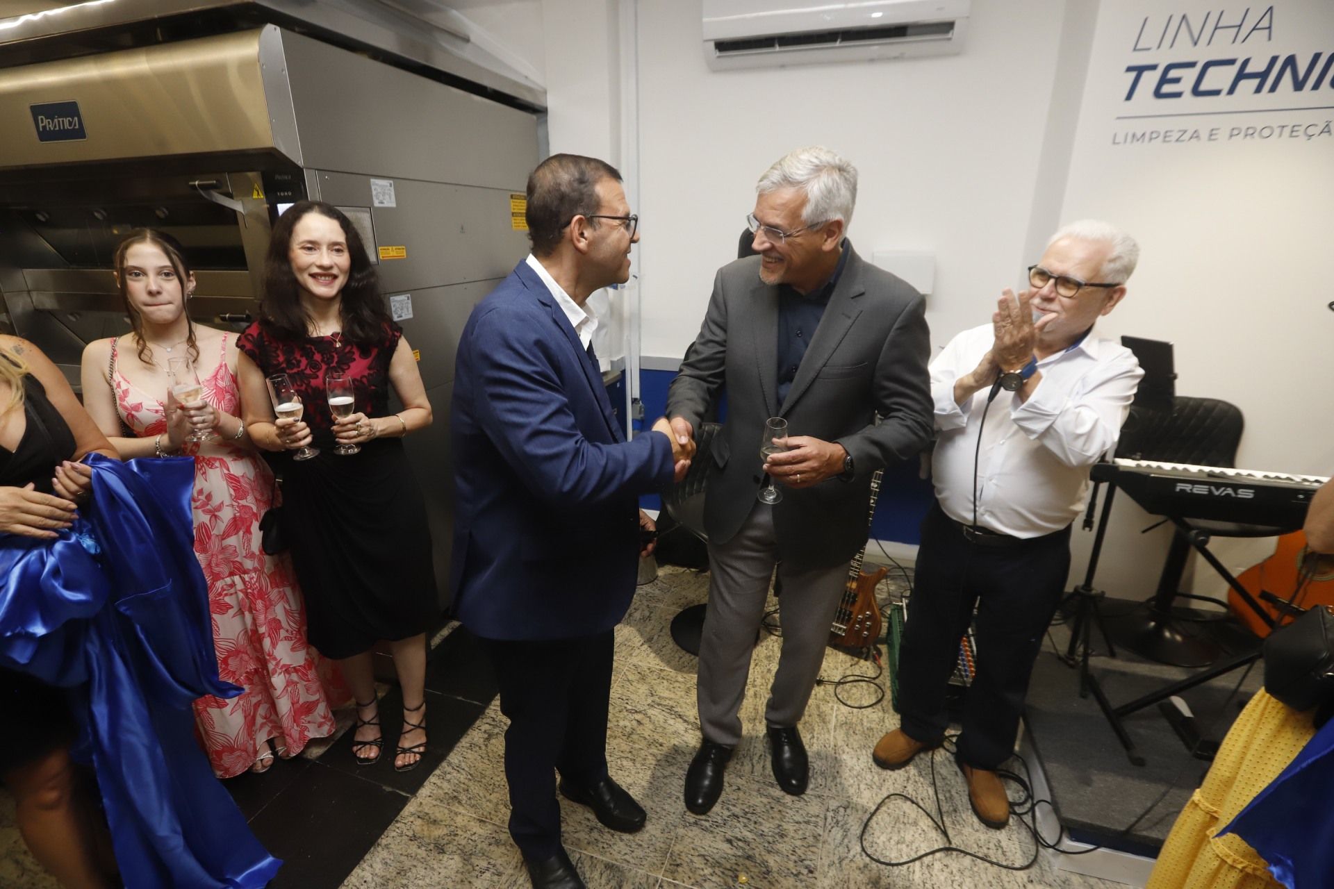 Dois homens apertando as mãos em um ambiente de negócios, outros aplaudem, uma fita é segurada e uma faixa diz 'Linha Techni'.