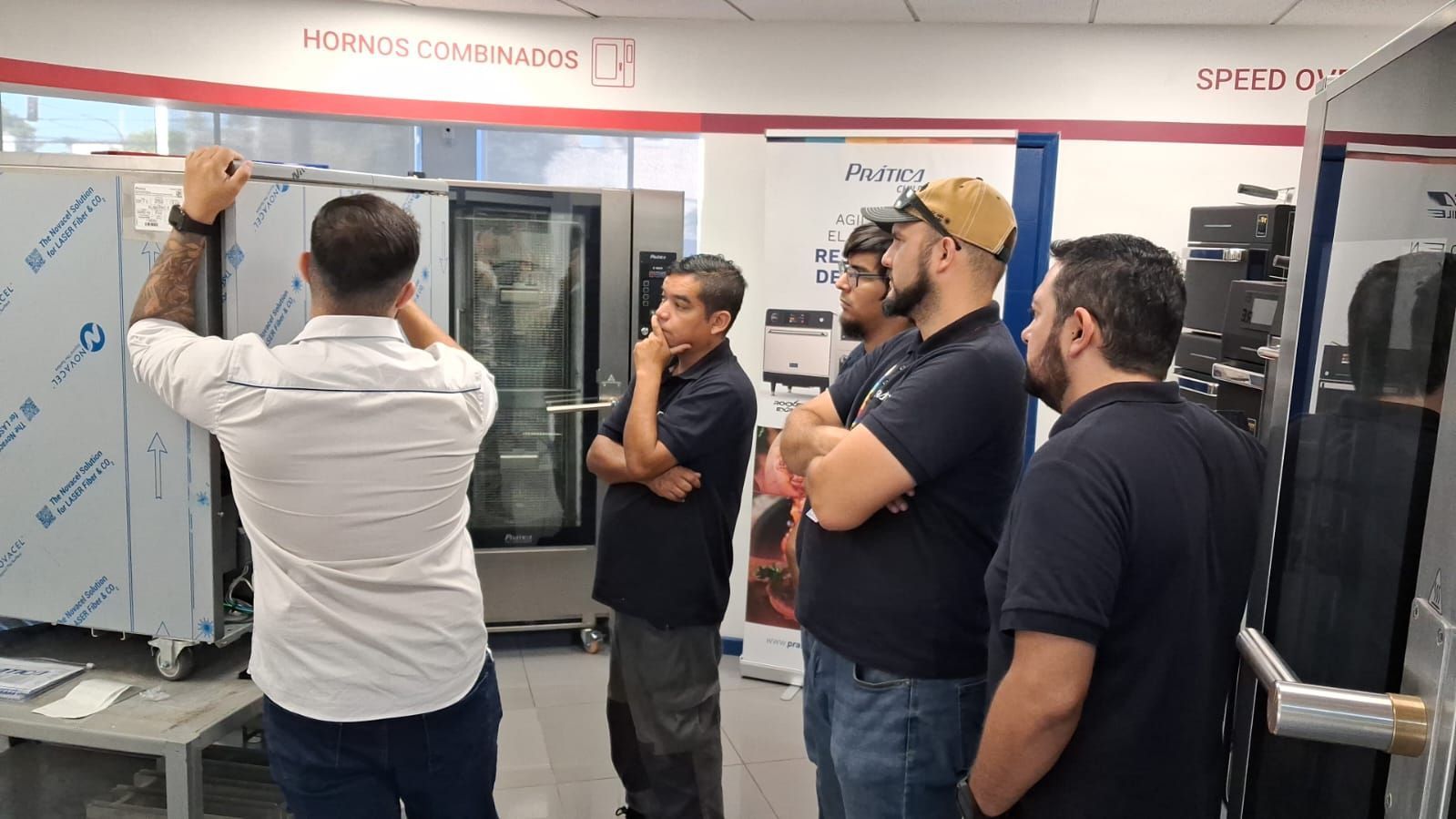 Grupo de homens observando uma demonstração de forno dentro de uma loja, um deles apontando.