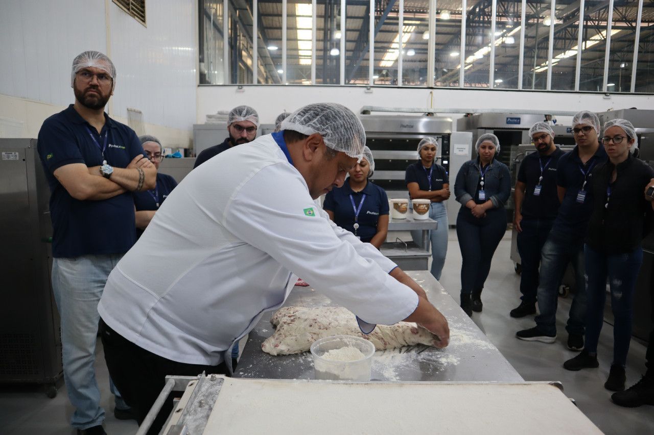 Um homem está amassando massa na cozinha enquanto um grupo de pessoas observa.