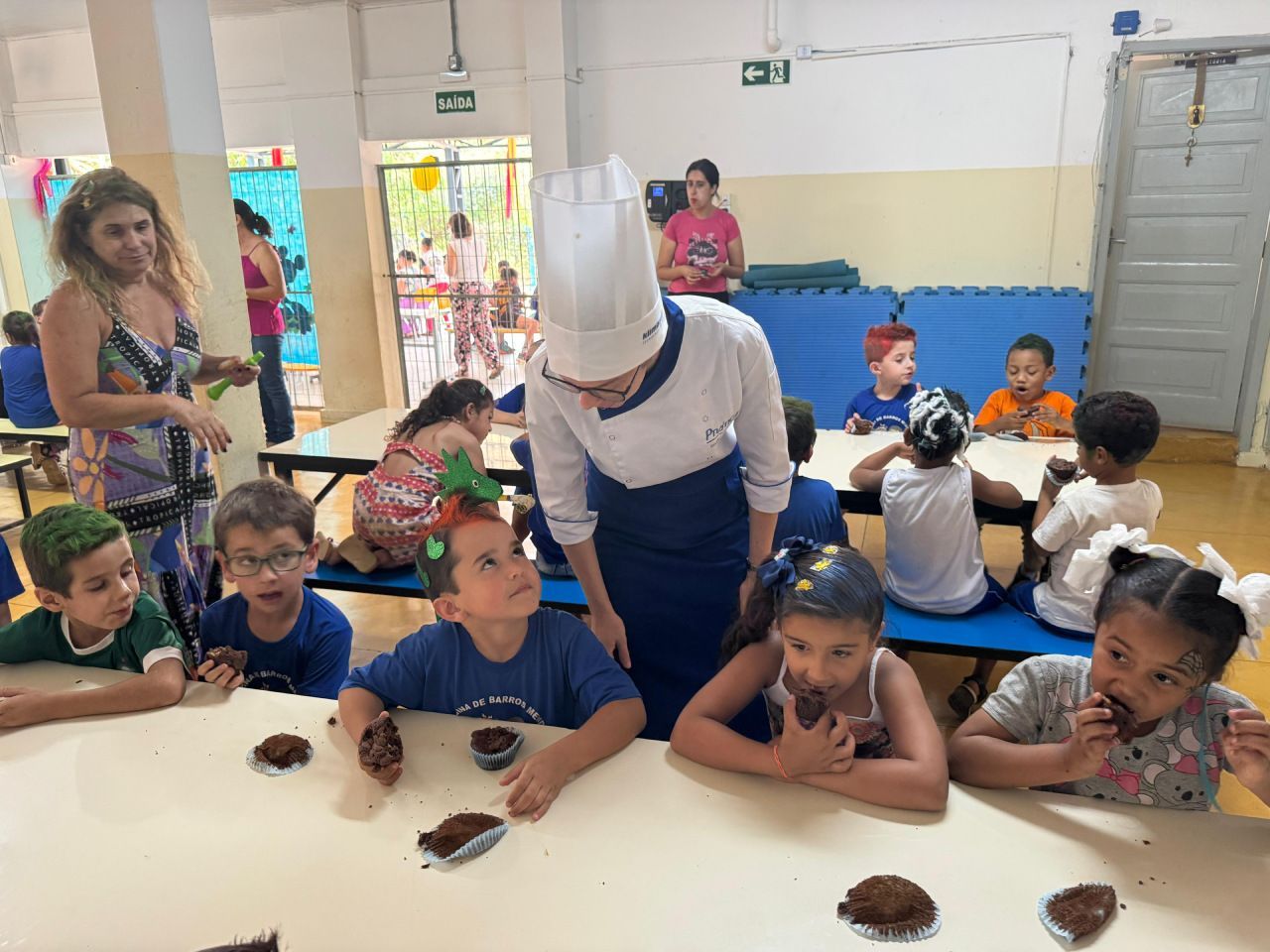 Um homem com chapéu de chef está conversando com um grupo de crianças em uma mesa.