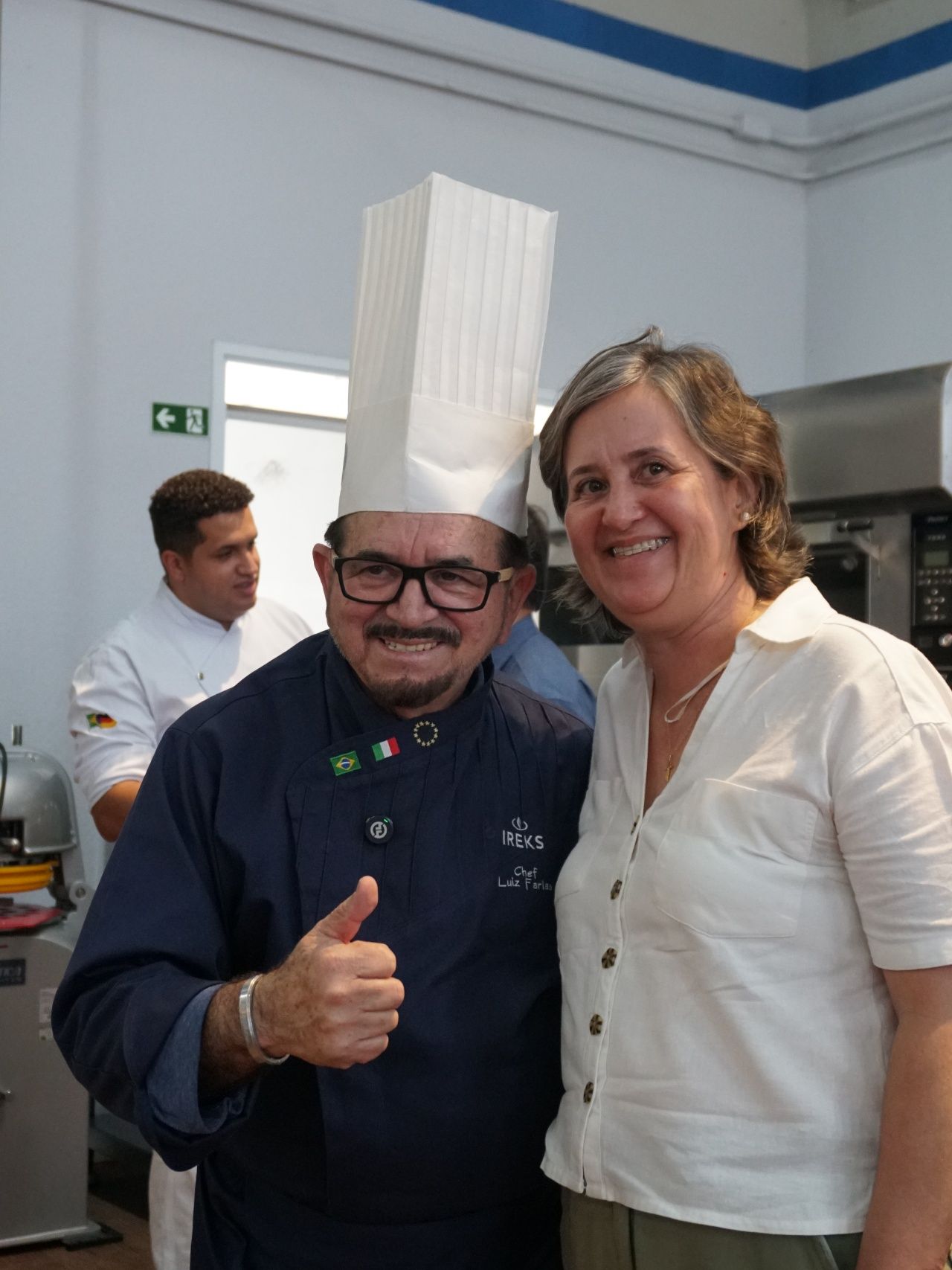 Chef de chapéu alto faz sinal de positivo e sorri para uma mulher na cozinha. Outro chef ao fundo.
