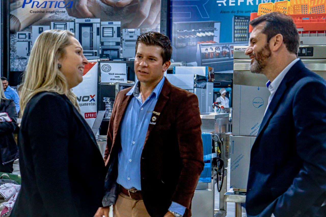 Um grupo de pessoas estão de pé, uma ao lado da outra, em uma sala, conversando entre si.