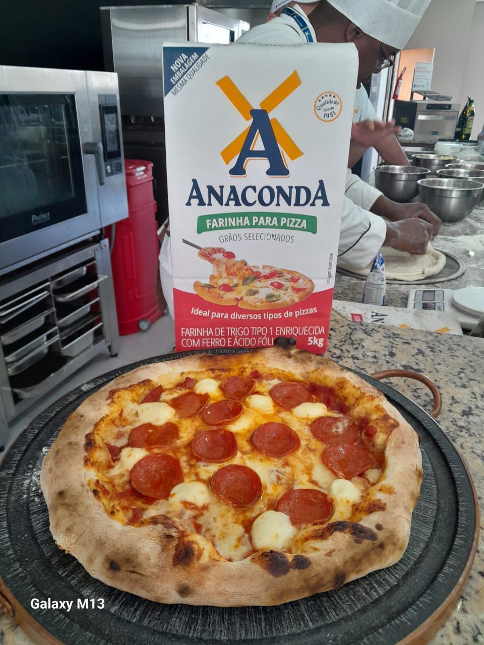 Uma pizza de calabresa está sobre uma mesa ao lado de uma caixa de massa de pizza anaconda.