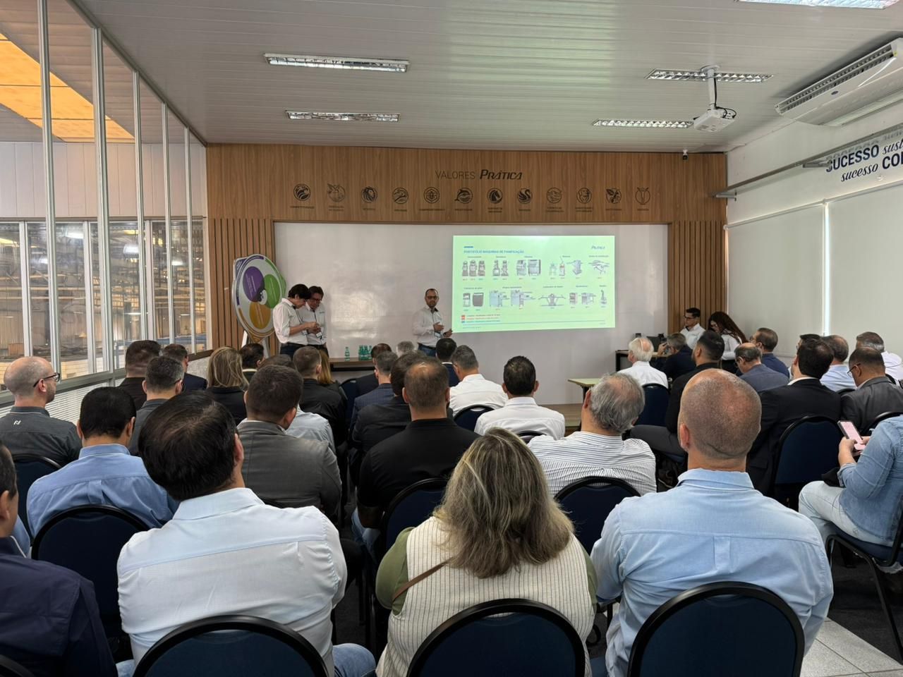 Apresentação em uma sala: palestrante, tela com diagrama, público sentado de frente.