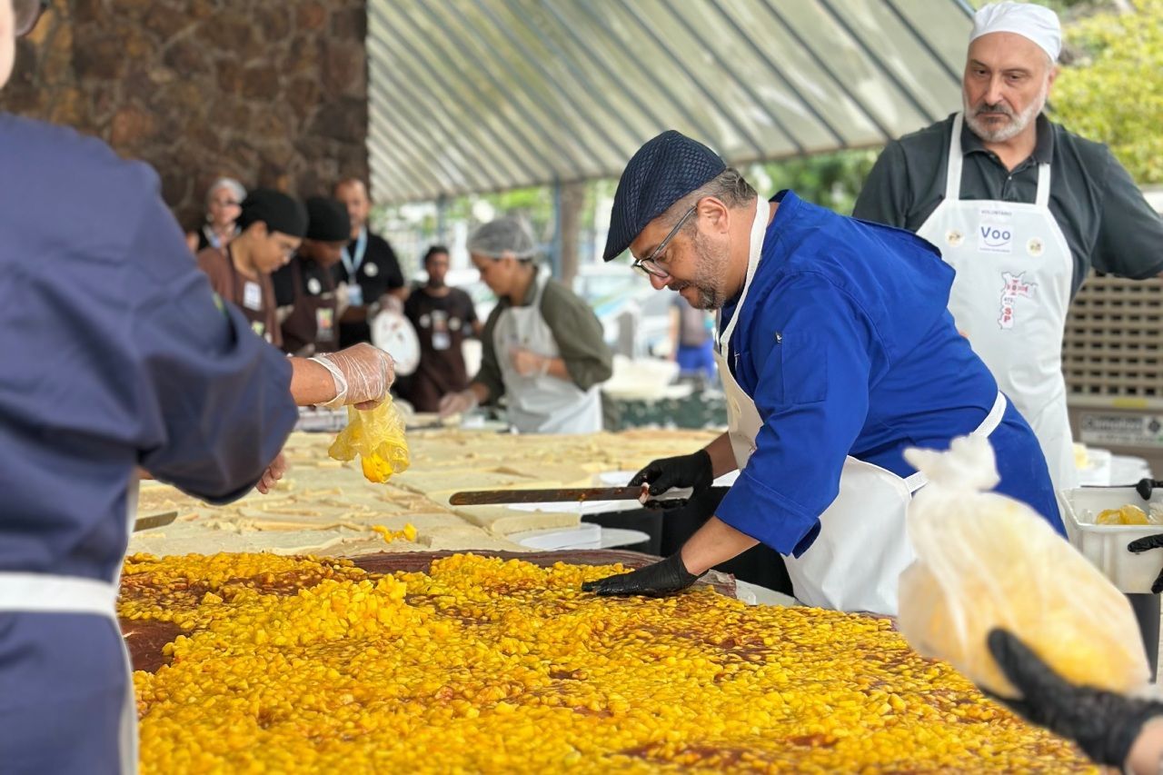 Chefs preparando paella gigante em uma grande mesa, ao ar livre. Pessoas de avental e chapéu, trabalhando com o arroz amarelo.