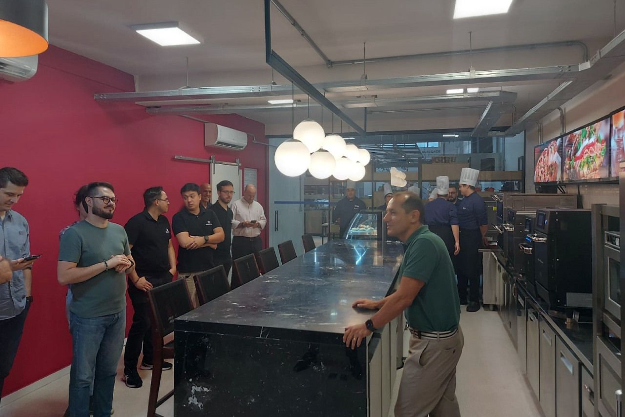 Um grupo de homens está em pé ao redor de uma longa mesa na cozinha.