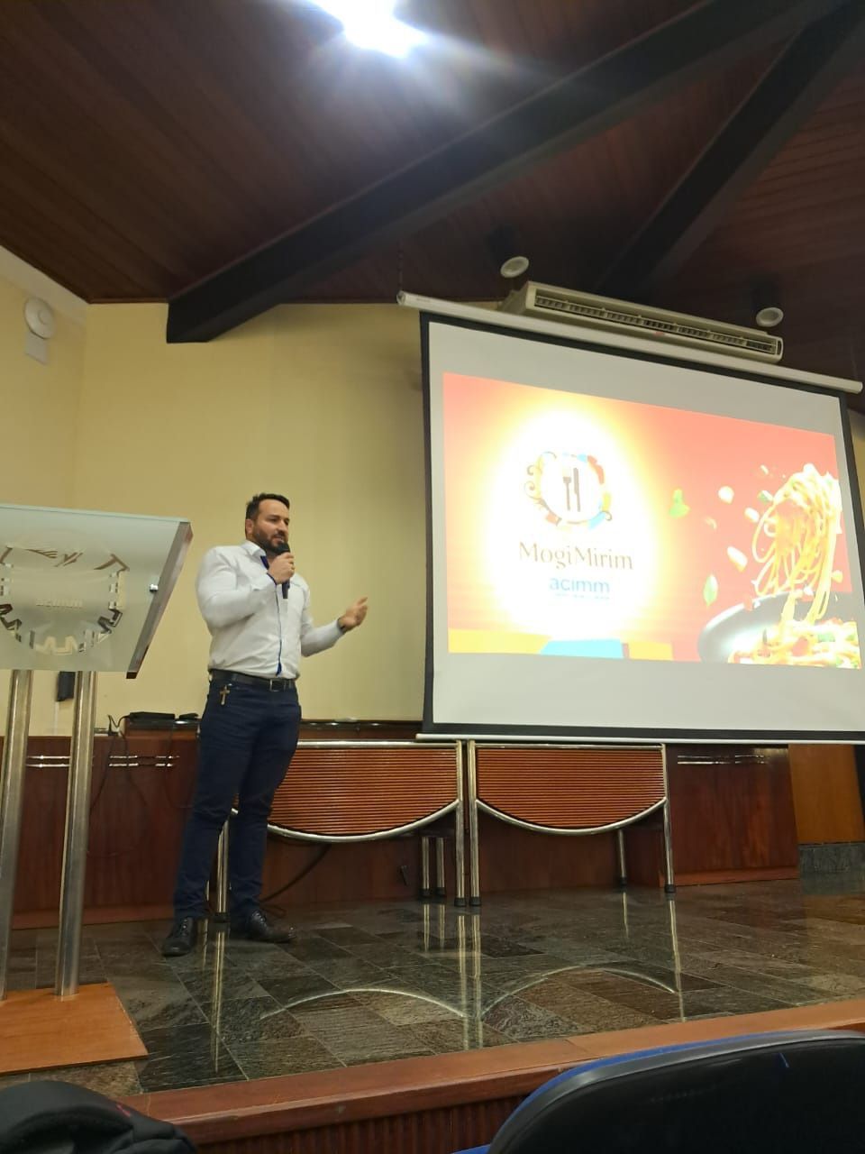 Homem fazendo apresentação em um pódio, em pé em um palco com uma tela de projetor em uma sala de aula.