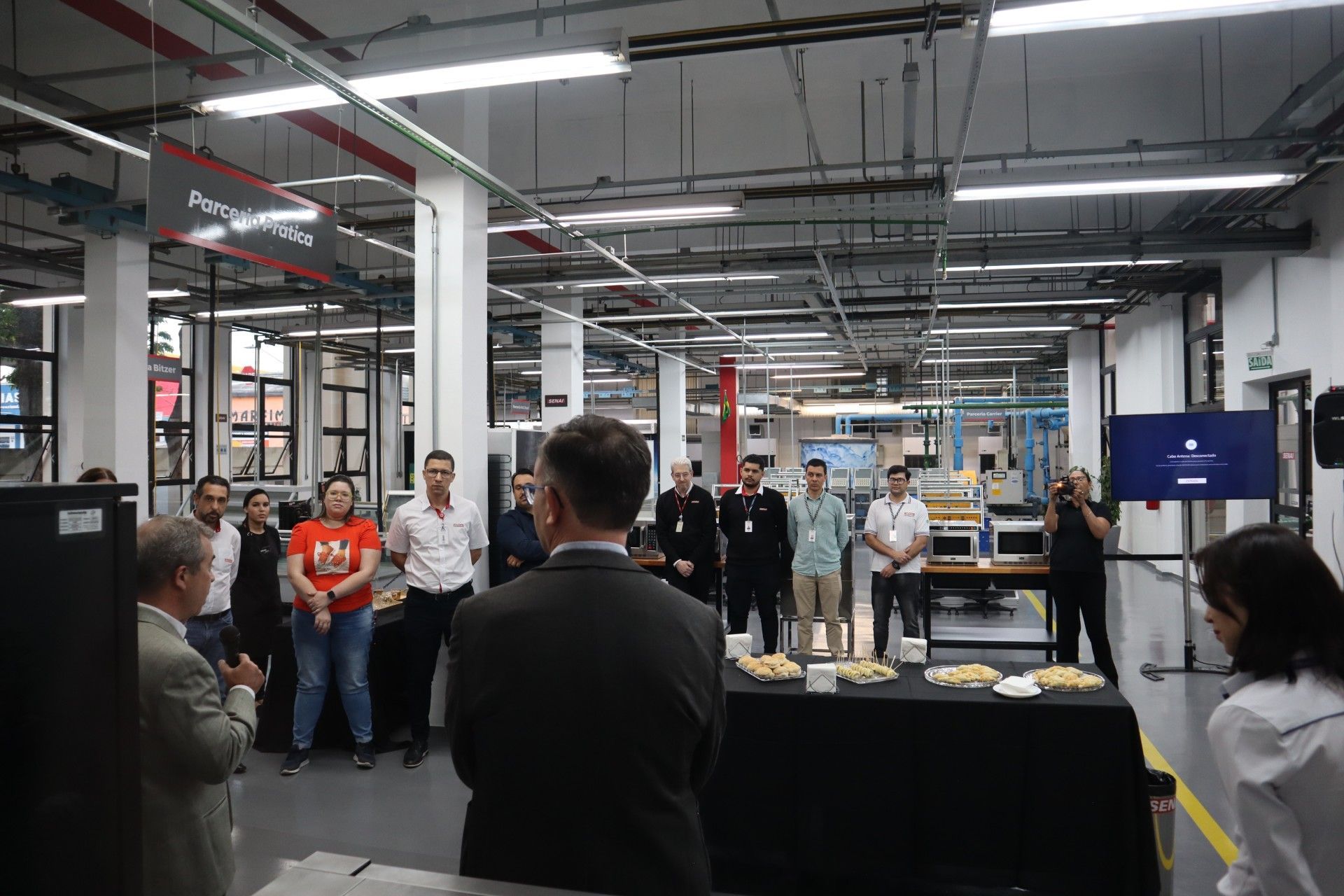 Pessoas reunidas em um espaço industrial, algumas olhando para a frente. Uma tela de apresentação é visível.