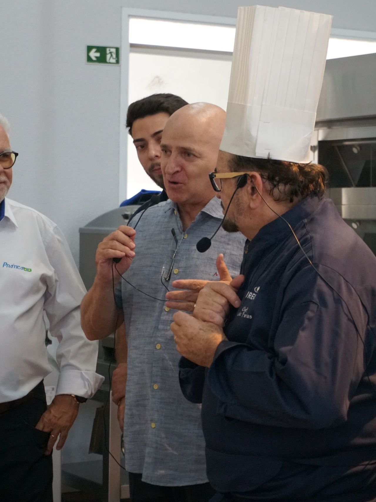 Chefs conversando em uma cozinha. Um homem com um chapéu alto de chef gesticula, enquanto outros escutam, alguns com microfones.