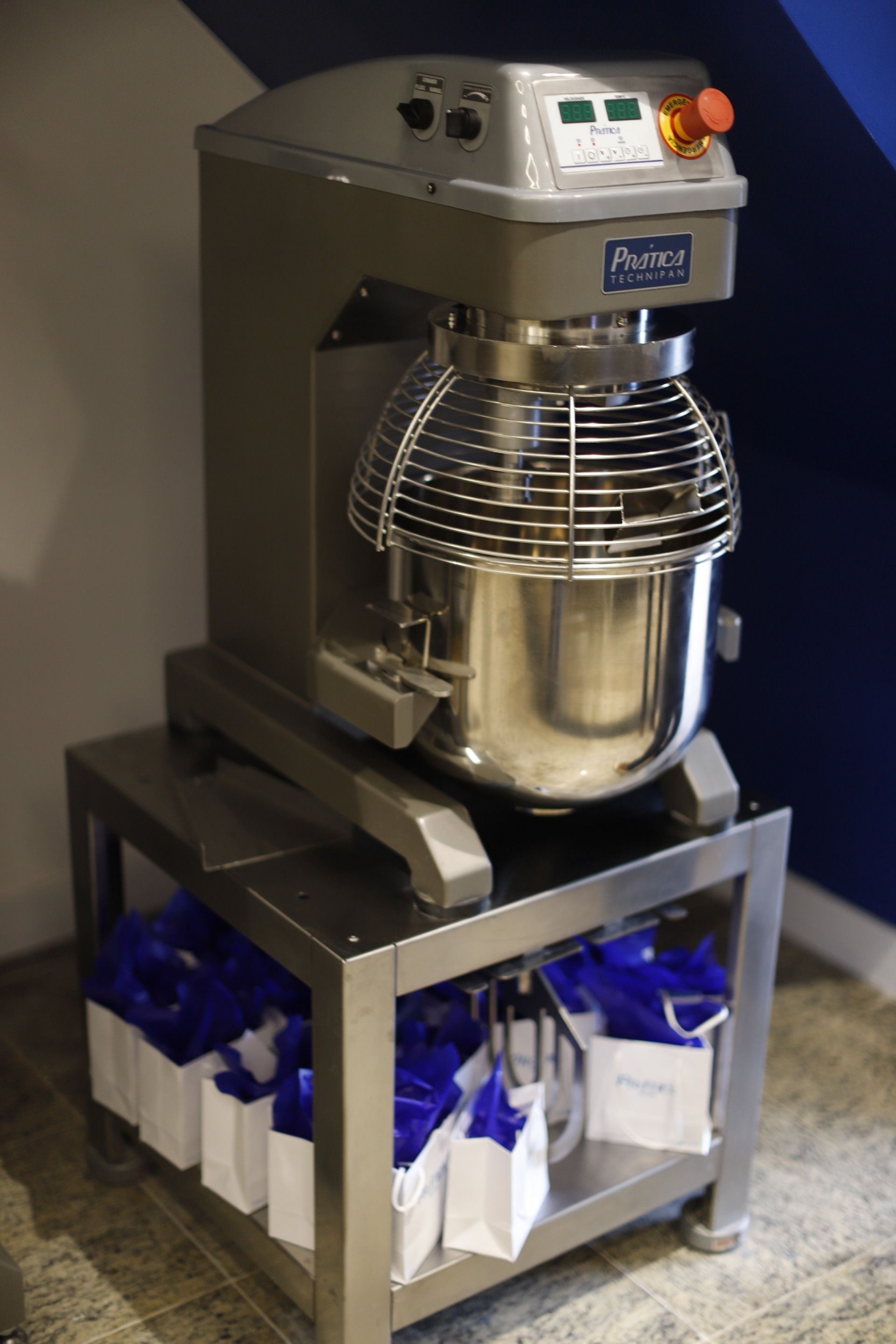 Grande batedeira comercial prateada com gaiola e tigela de metal, sobre uma prateleira de aço inoxidável com sacolas de presente embrulhadas em azul abaixo.