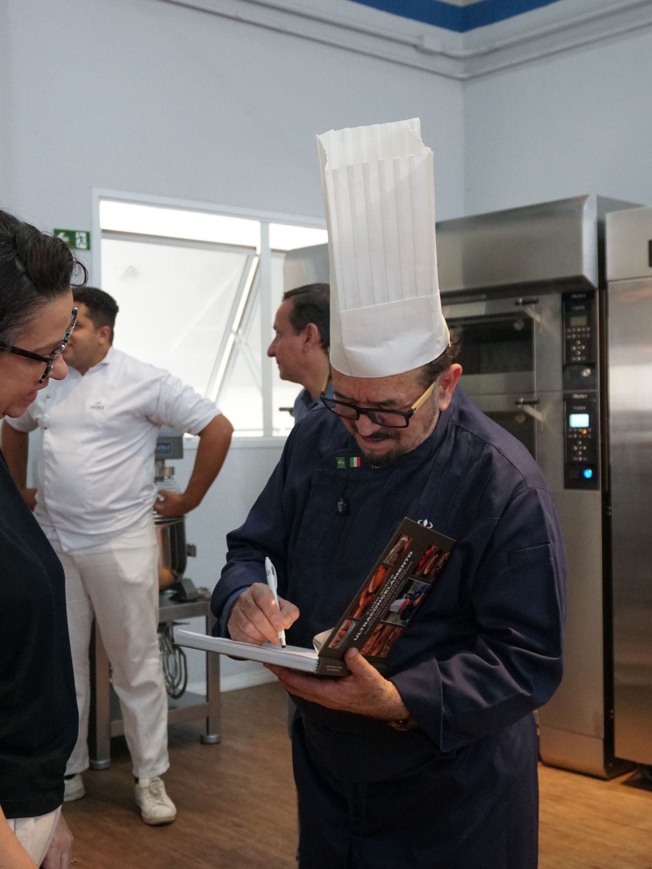 Chef de chapéu alto assinando papéis na cozinha; outros observam.