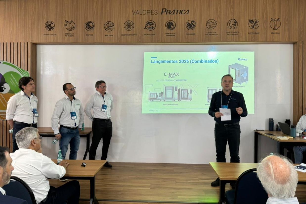 Uma apresentação em uma sala de conferências; quatro homens ficam perto de uma tela e falam para o público.