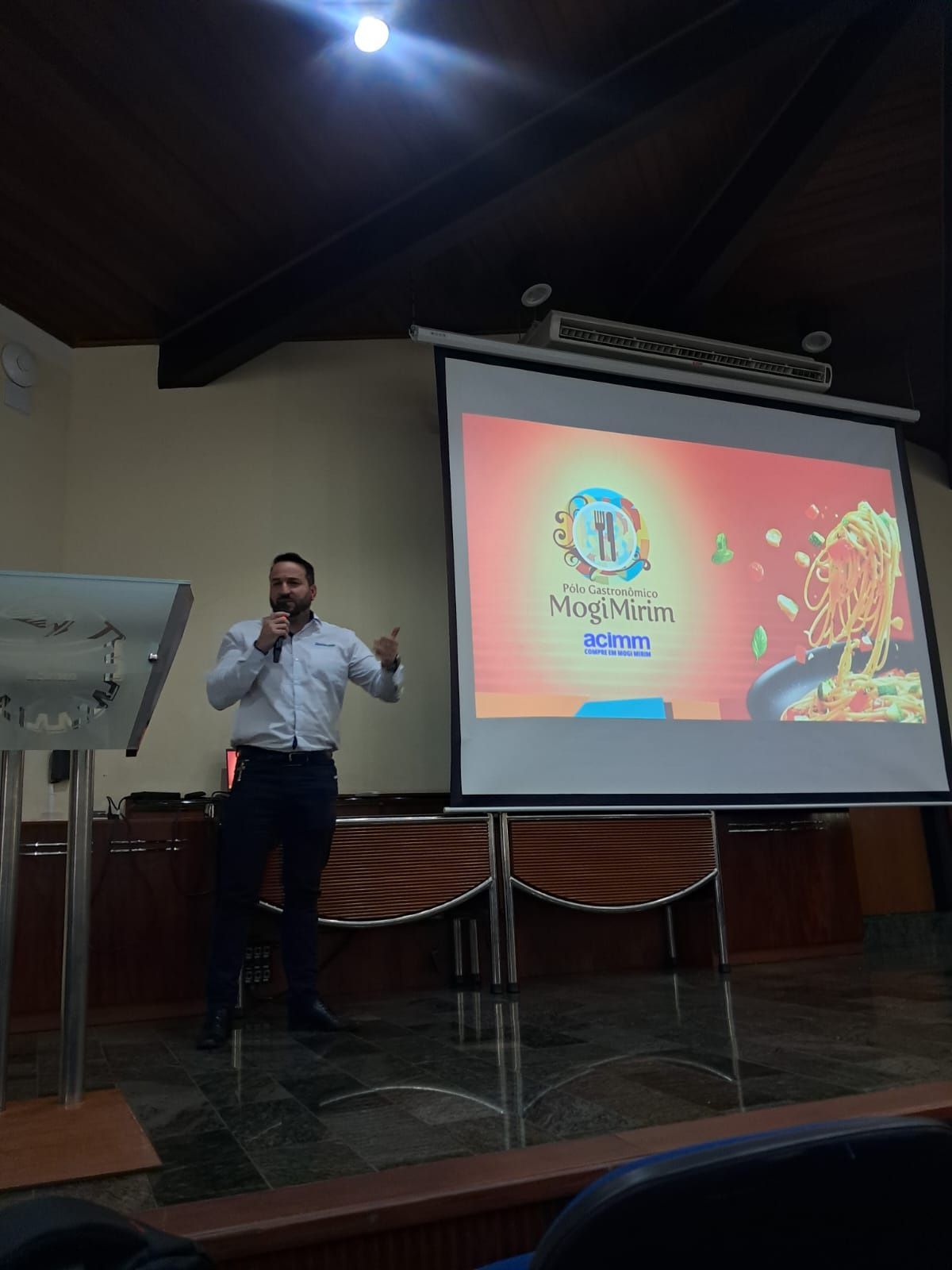 Homem fazendo uma apresentação em frente a uma tela, em ambiente interno. A tela exibe o logotipo e as imagens. O homem gesticula, vestindo uma camisa clara e calças escuras.